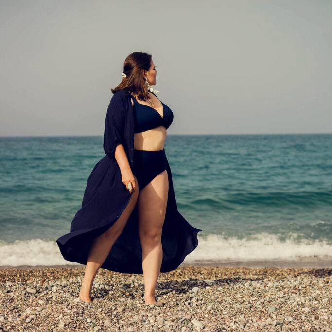 Attractive busty curvy woman in a blue swimsuit resting on the beach. Stylish accessories, fringe, fashion for plus size, beautuful sea. Bodypositive, natural authentic beauty, resort, summer vacation. Copy space.